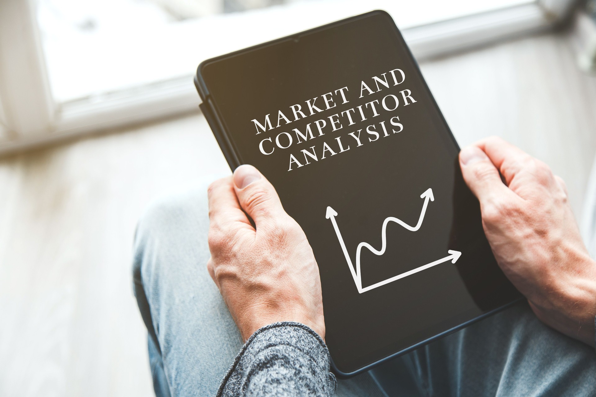Close-up of a man holding a tablet with the words text Market and Competitor Analysis and a rising line graph on the screen, concept of strategic planning and market research